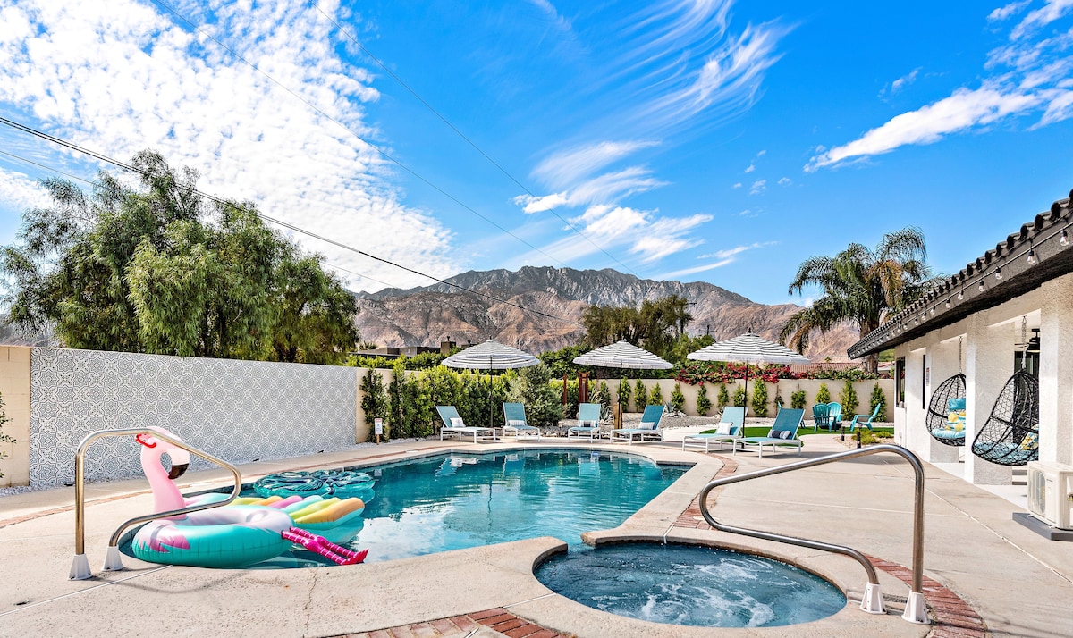 A resort-style backyard features a large swimming pool with colorful floats resting on the water. Umbrellas provide shaded seating areas, while lounge chairs line the pool's edge. The stunning San Jacinto mountains serve as a backdrop, enhancing the outdoor space's tranquil atmosphere.