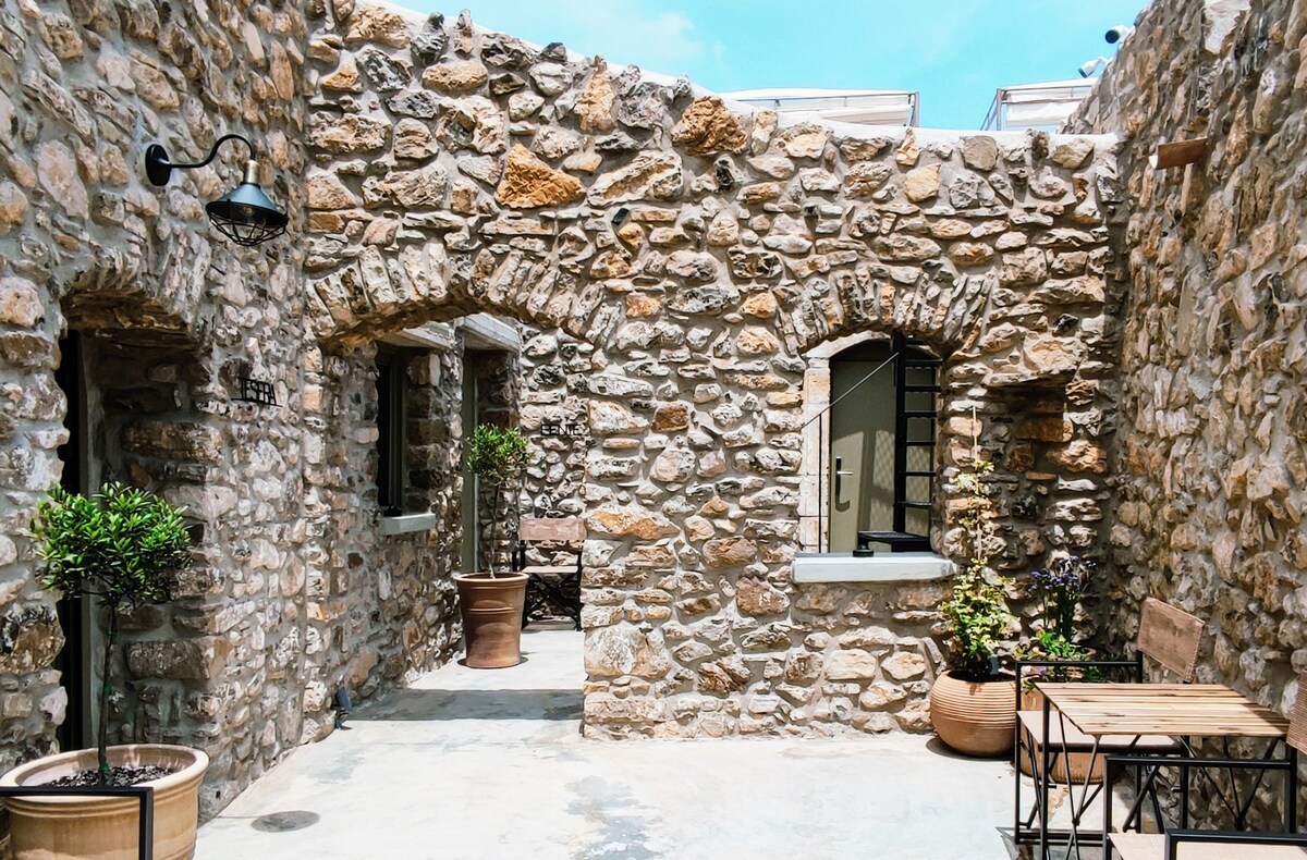 Exposed stone walls define the charming courtyard, showcasing a blend of natural textures. Potted plants add greenery, while simple wooden furniture provides inviting seating areas. Arched openings frame views of the exterior, enhancing the rustic atmosphere of the restored Cretan property.