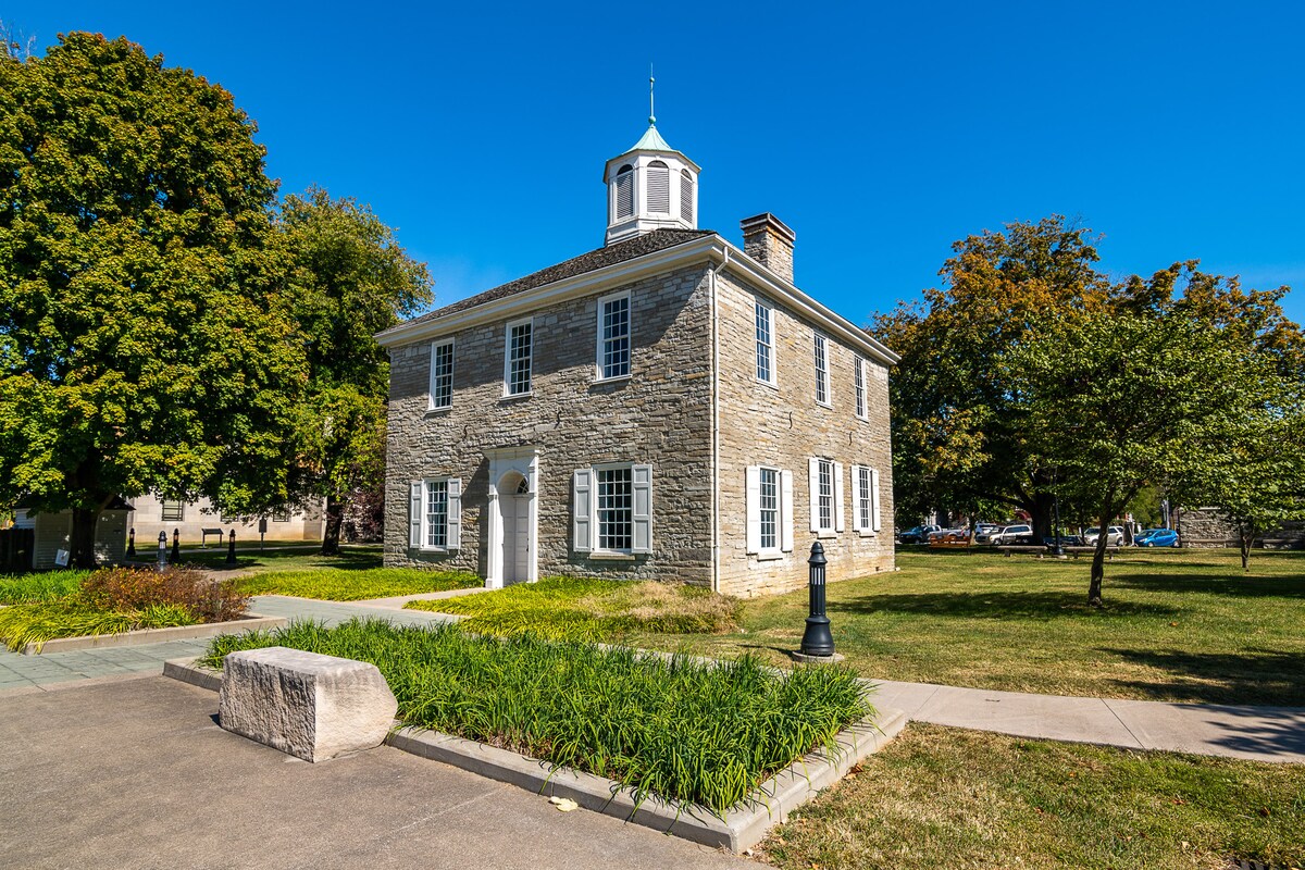 A historic stone building is showcased with a charming cupola atop its roof. Lush greenery surrounds the structure, which is complemented by well-maintained pathways and landscaped areas. Large windows offer a view of the inviting interior, while trees provide shade and enhance the peaceful atmosphere.