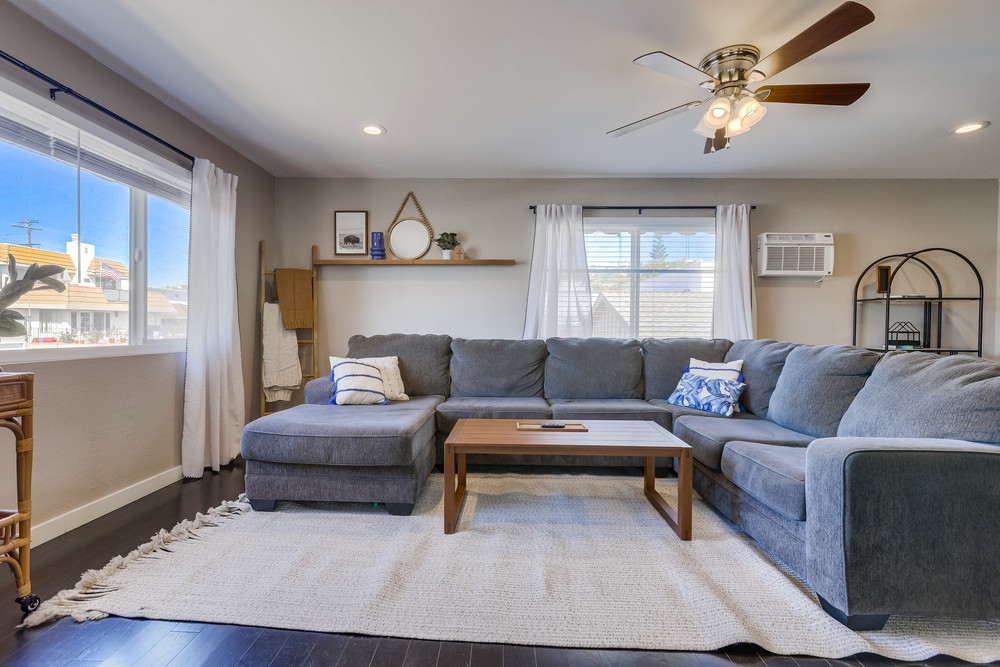 A spacious living area is furnished with a large sectional couch and a wooden coffee table. Natural light enters through a wide window dressed with light curtains. A ceiling fan hangs overhead, and decorative shelves are mounted on the wall.
