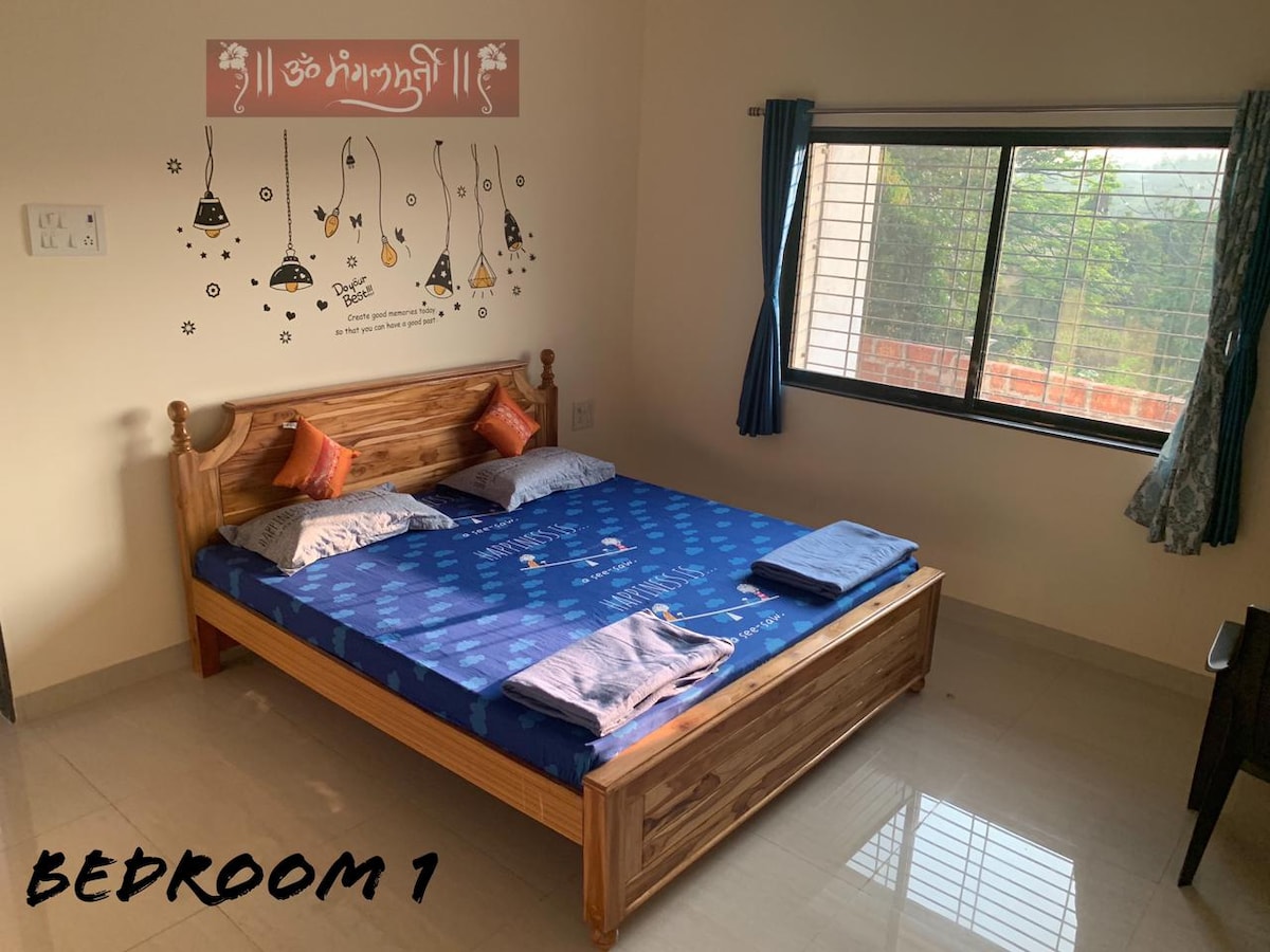 A welcoming bedroom features a king-size bed dressed with blue bedding and two pillows. A window on one wall allows natural light to enter, offering a view of the greenery outside. A decorative wall decal adds a personal touch. Soft towels are neatly placed on the bed.