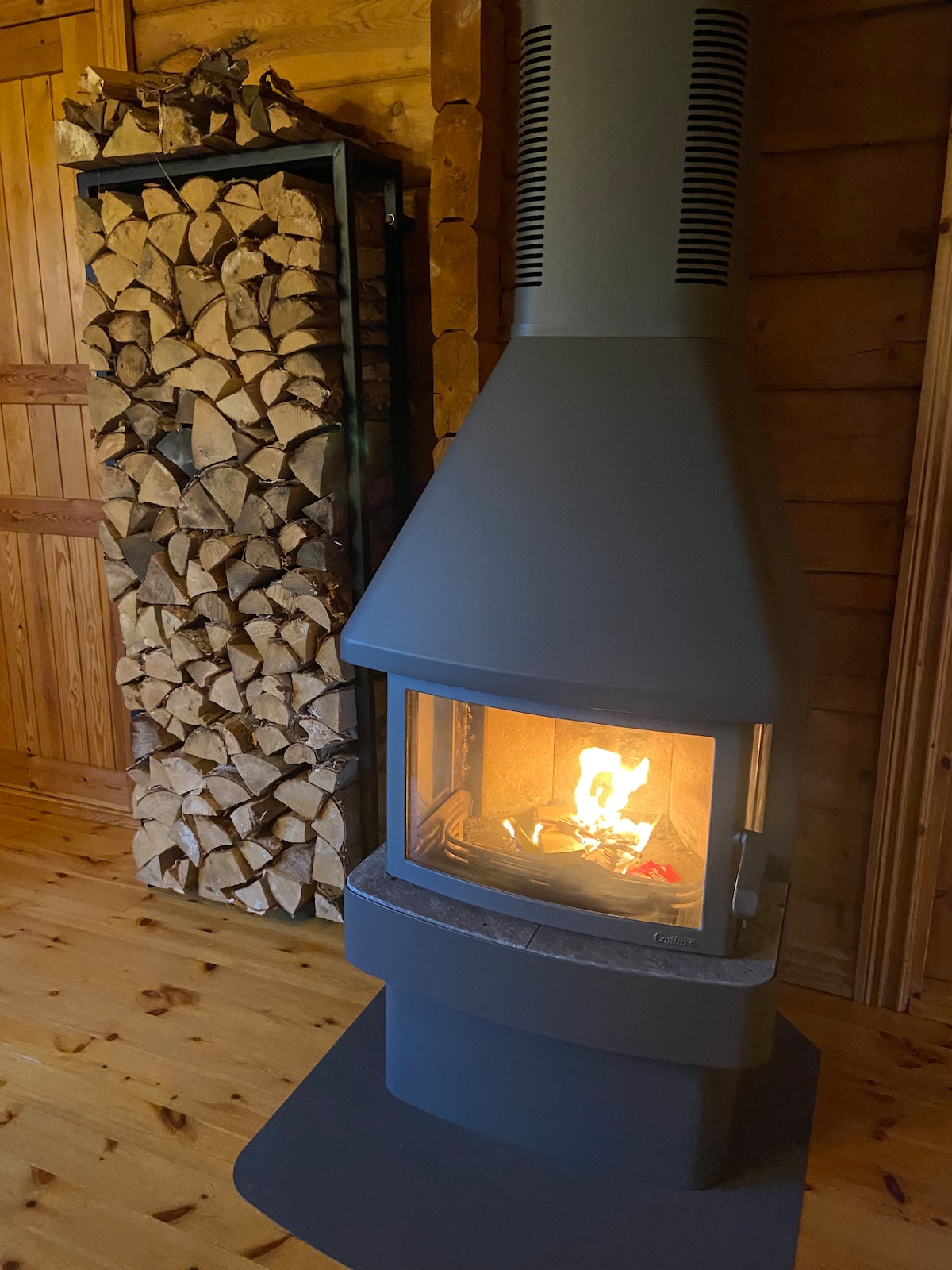 A modern wood stove is positioned in a corner, providing warmth with an inviting flame visible through the glass front. Stacked firewood is neatly arranged in a vertical rack nearby, complementing the rustic wooden interiors and enhancing the cozy ambiance.