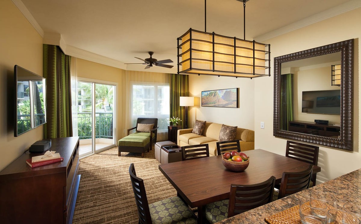 A spacious living area is highlighted by a large window offering natural light. A comfortable sofa and armchair complement a wooden dining table with chairs. A stylish lantern light fixture hangs above, while a flat-screen TV is mounted on the wall, providing modern entertainment options.