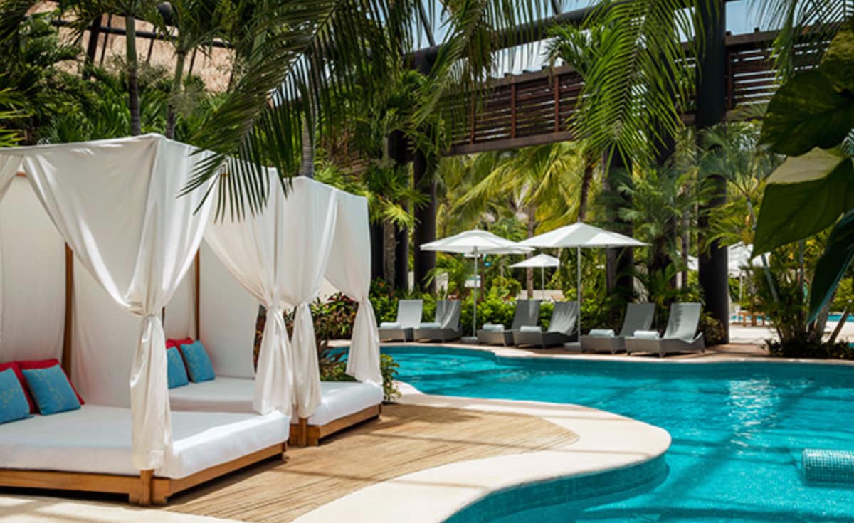 Private cabanas are situated beside a clear pool, surrounded by lush tropical plants. Sun loungers are arranged nearby under shaded umbrellas, offering an inviting area for relaxation. The bright sunlight creates a serene environment, highlighting the resort's tranquil setting.