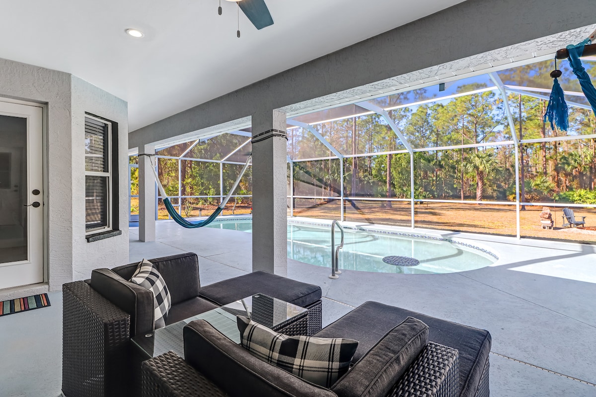An outdoor seating area is furnished with a dark wicker couch featuring a patterned cushion. A spacious saltwater pool is visible through a screened enclosure, surrounded by a tranquil landscape. Two hammocks and lounge chairs are positioned for relaxation.