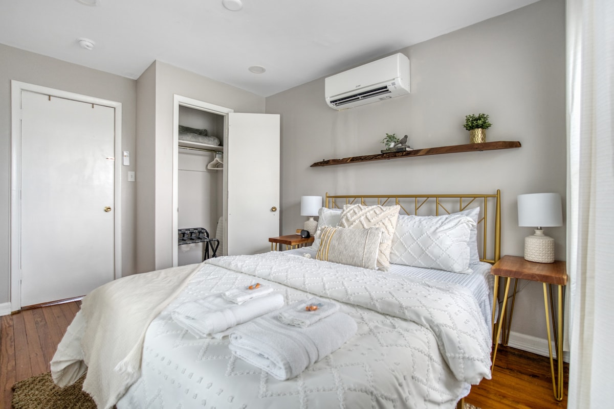 A light-filled bedroom features a spacious queen bed adorned with crisp linens and two folded towels atop. Nightstands with lamps sit on either side of the bed. A closet space is visible, along with a cozy air conditioning unit mounted above the bed.