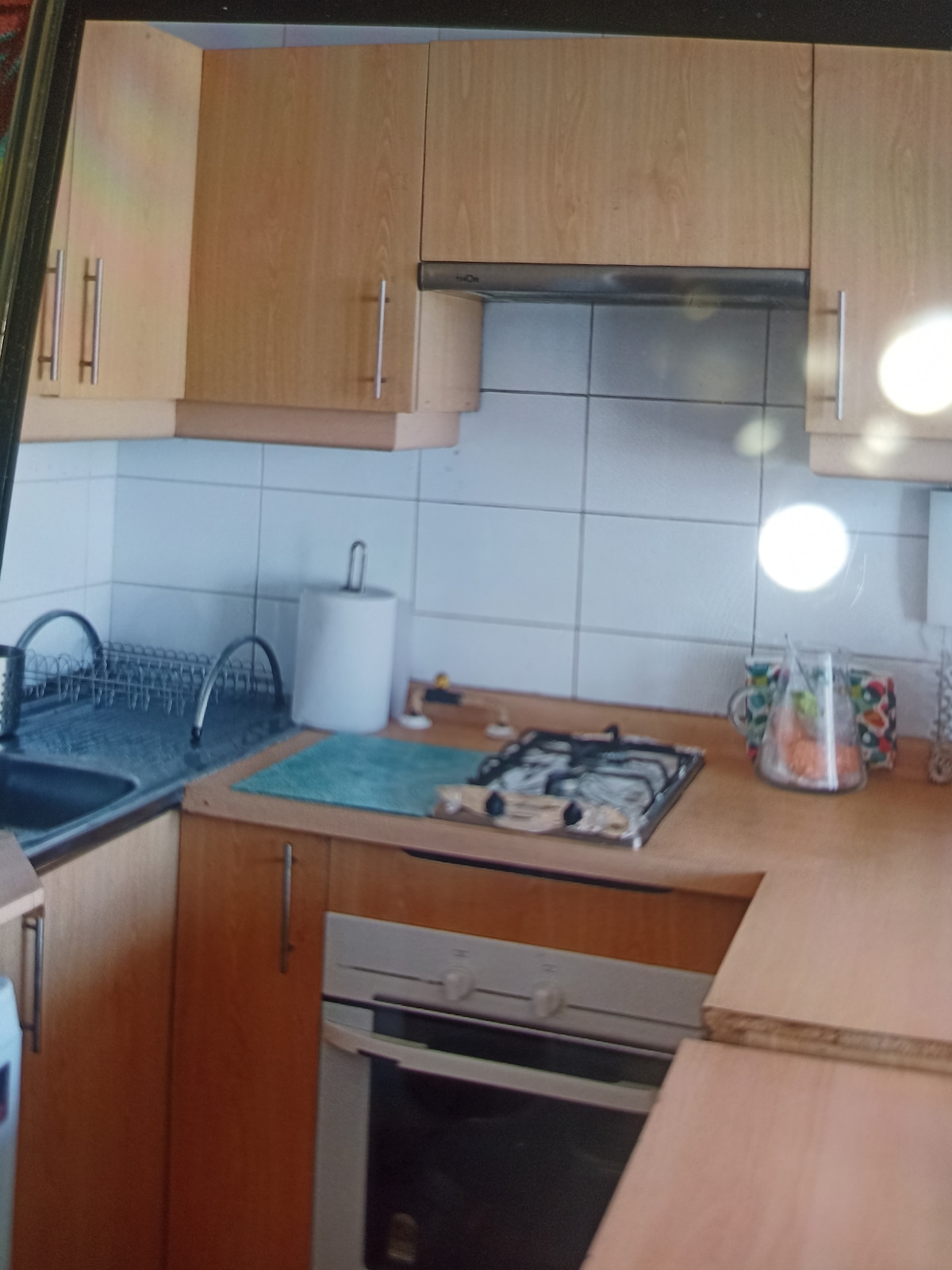 An equipped kitchen features wooden cabinets and a countertop space. A gas stove is located next to a sink, while a white countertop appliance is placed for convenience. A colorful bowl holds various fruits, adding a touch of brightness to the area.