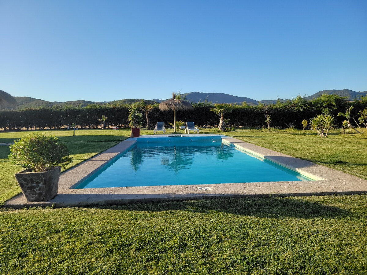 A tranquil swimming pool is surrounded by neatly manicured grass and framed by lush greenery. Two lounge chairs are positioned beside the pool, inviting relaxation. The distant hills are visible under a clear blue sky, enhancing the serene outdoor setting.