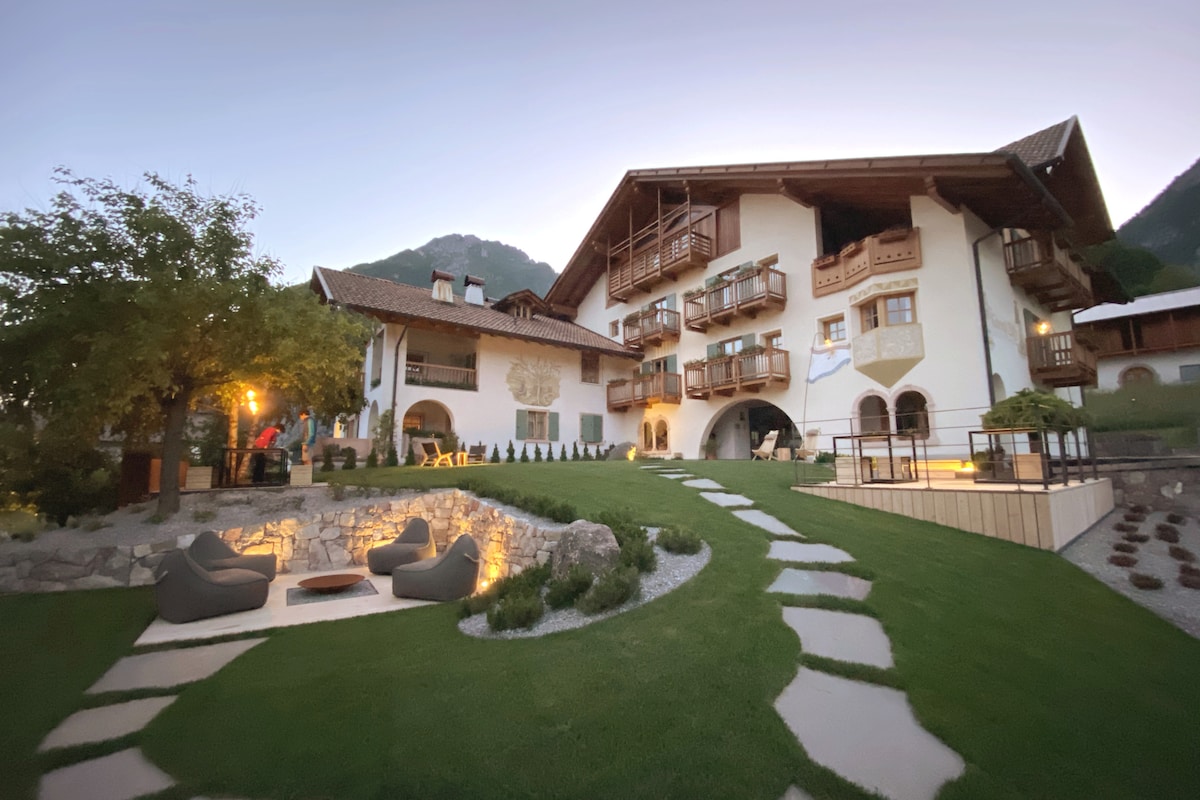 The exterior of the structure features a traditional Trentino design with wooden balconies and archways. A manicured garden is visible in the foreground, highlighted by soft outdoor lighting. Comfortable seating with smooth gravel pathways leads to the entrance, surrounded by lush greenery and distant mountainous scenery.