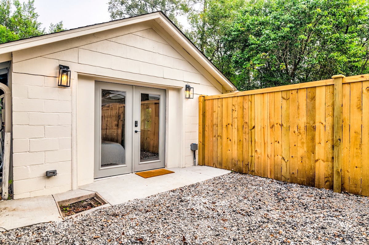 An exterior view of the detached guesthouse is shown, featuring double glass doors that open into the studio space. A wooden fence provides privacy, while a gravel pathway leads to the entrance, surrounded by greenery.