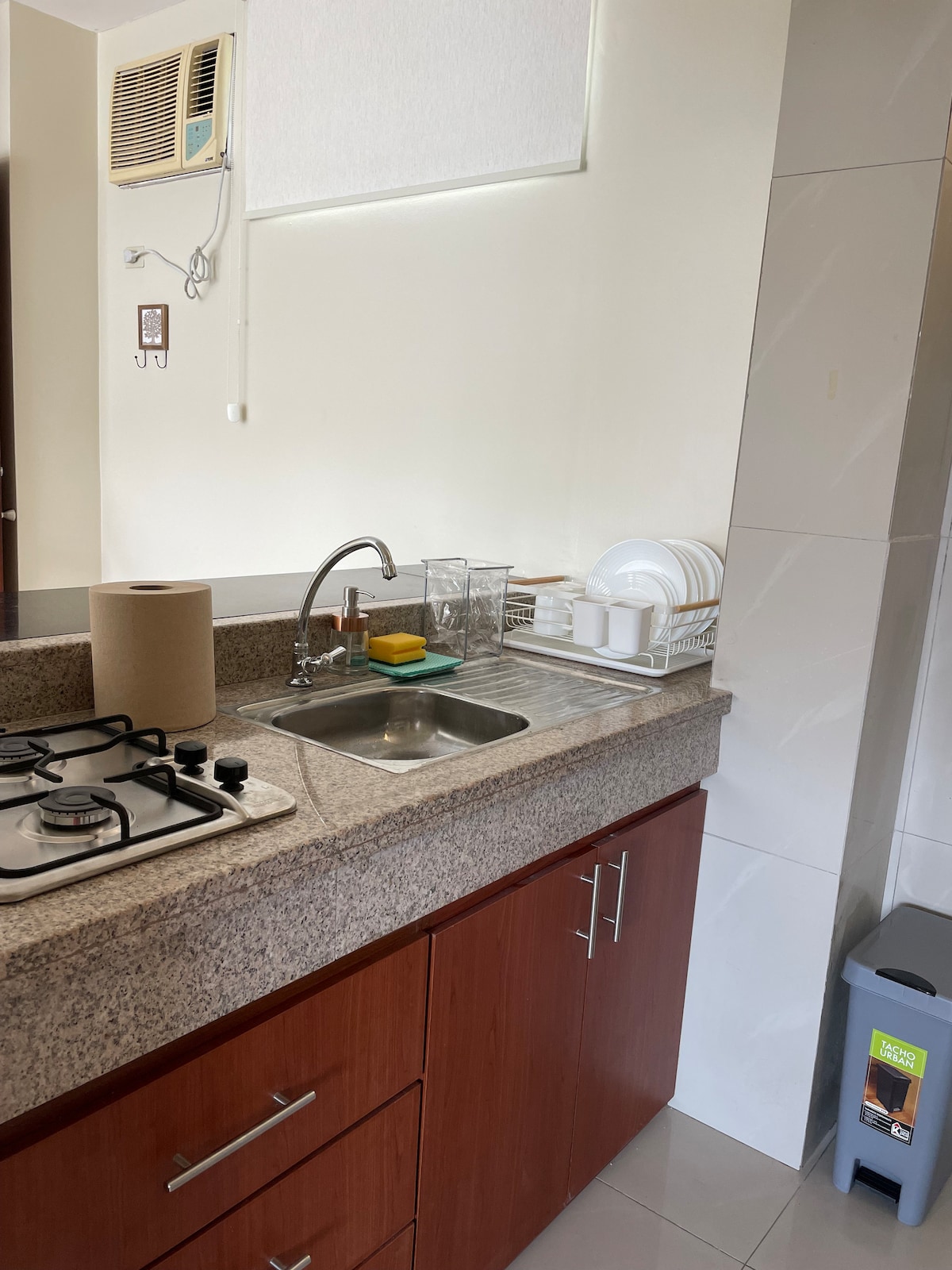 The kitchen area is equipped with a countertop featuring a sink and a gas stove. Clean dishes and glassware are arranged on a nearby drying rack. A roll of paper towels sits on the counter, alongside a trash can positioned on the floor.