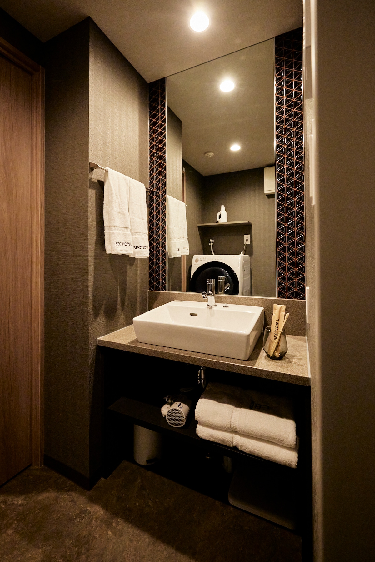 A compact bathroom features a modern sink positioned on a stone countertop. Above, a wall-mounted mirror reflects the ambient lighting. Clean white towels hang neatly on a rack, and toiletries are displayed beside a washing machine. Shelves below hold additional towels and supplies.