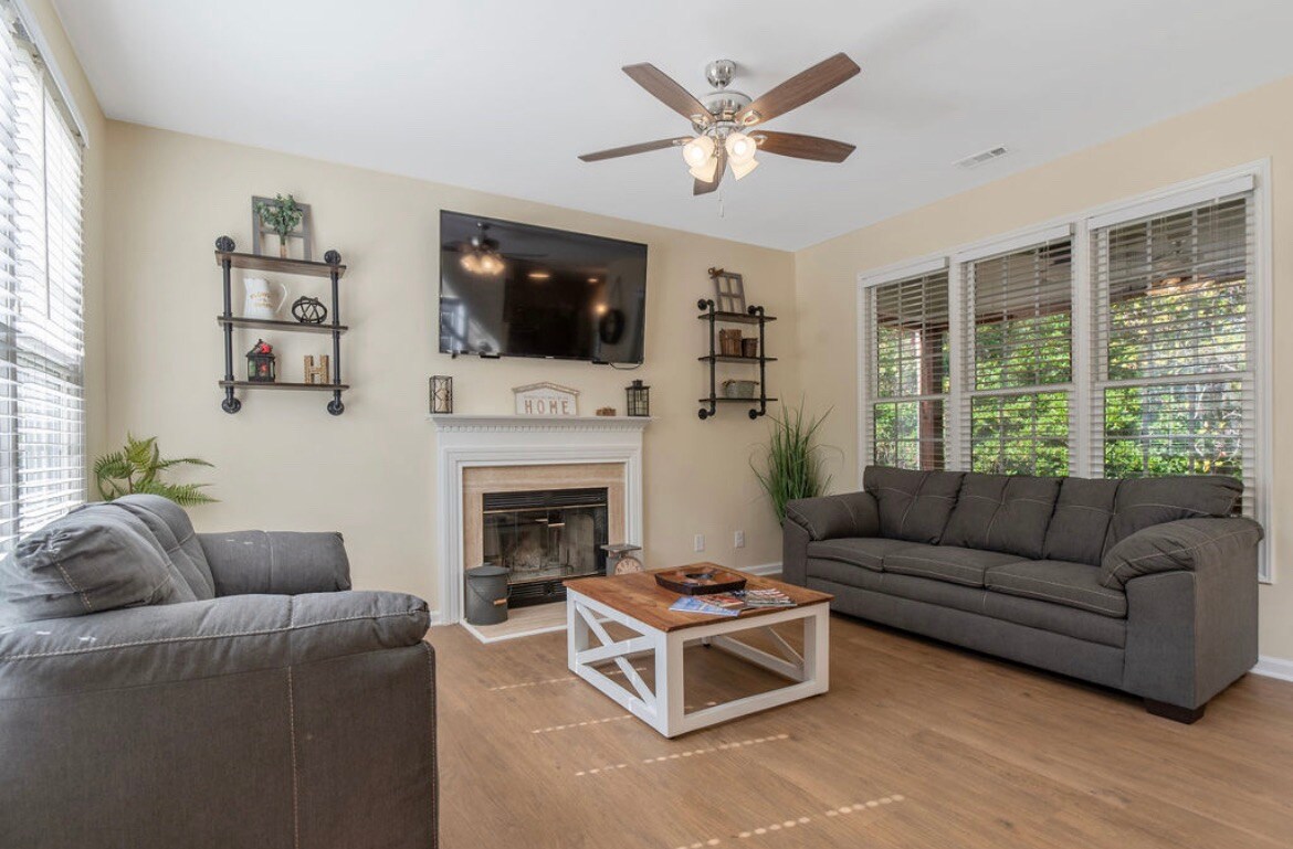 A cozy living area features comfortable seating, including a dark gray sofa and loveseat. A coffee table sits in the center, complemented by a decorative arrangement. Natural light filters through multiple windows, providing a bright and welcoming space, with a ceiling fan overhead for added comfort.
