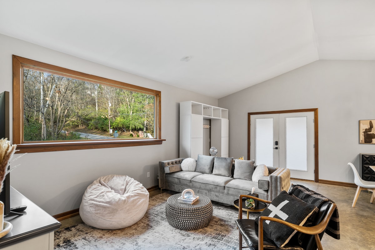 A spacious living area features a large window that frames a view of greenery outside. A comfortable sectional sofa and a soft bean bag are positioned around a low, decorative table. Bright walls and natural light contribute to a welcoming ambience.