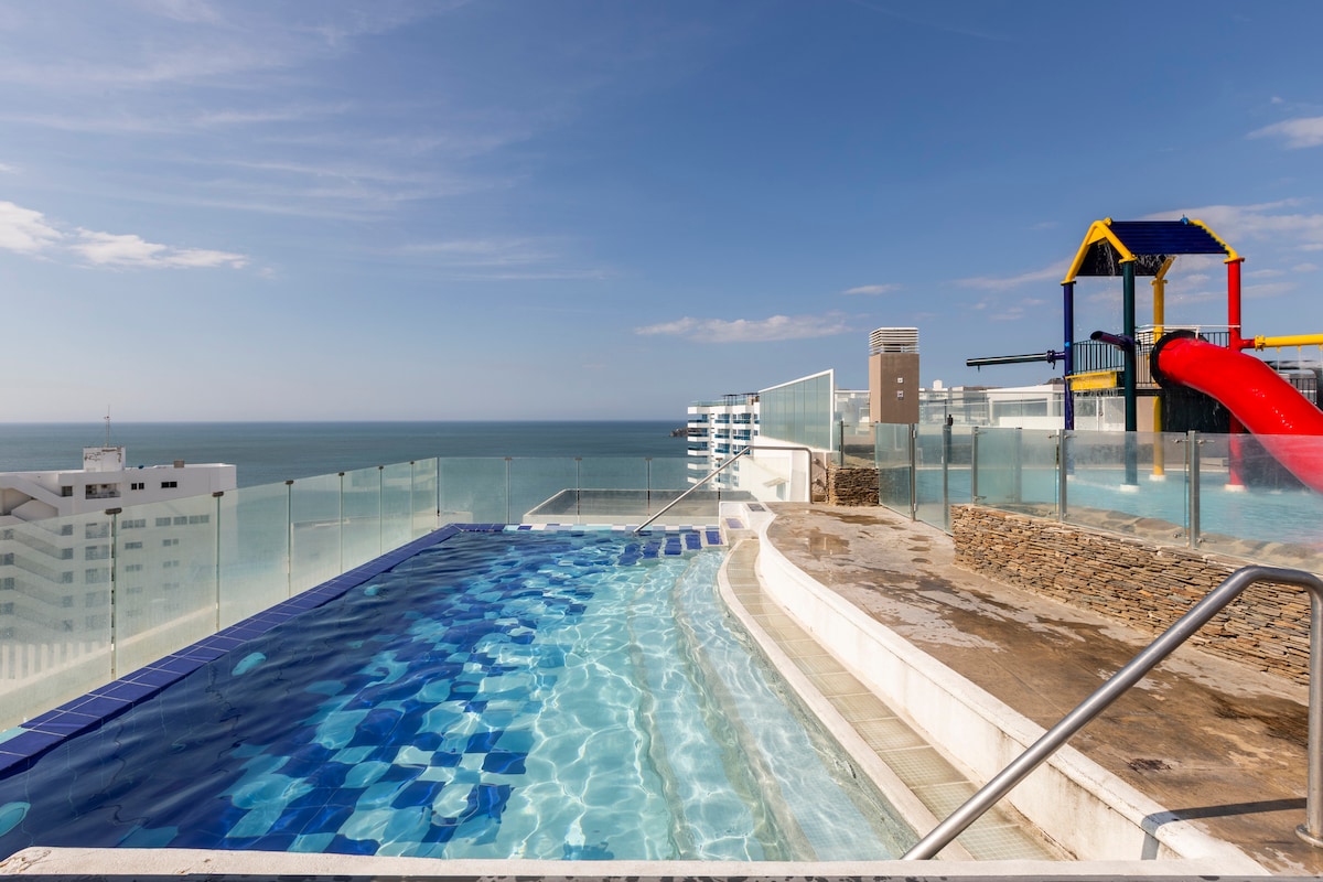 A rooftop pool with a shallow entry extends along the edge, offering an unobstructed view of the sea. A colorful playground structure sits nearby, featuring a slide and climbing frame. Glass railings provide safety while enhancing the open feel of the space.
