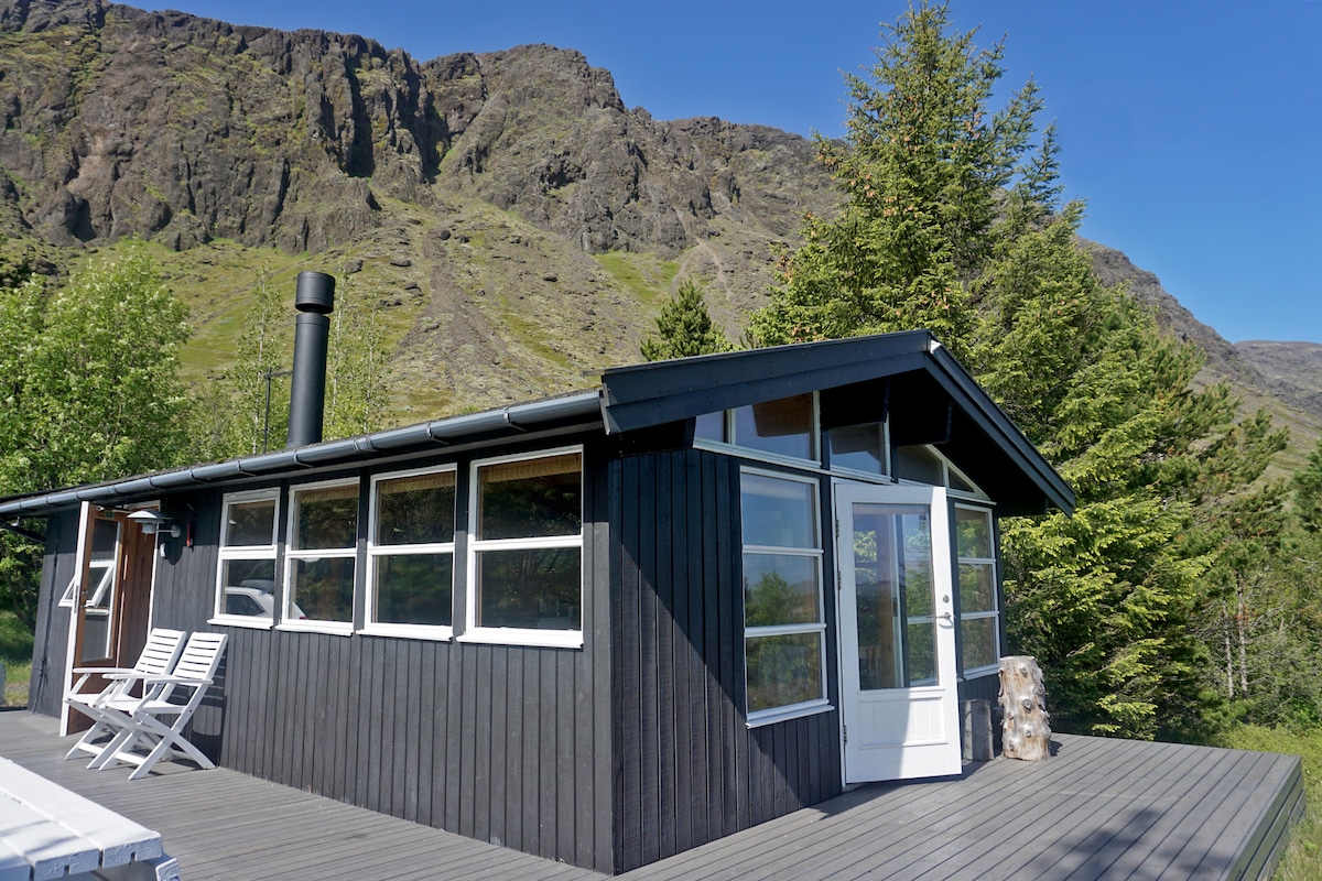 A modern cottage is situated against a backdrop of rugged mountains, featuring large windows that invite natural light. A sleek chimney rises from the roof, and a spacious deck presents multiple lounge chairs, surrounded by lush greenery and trees.
