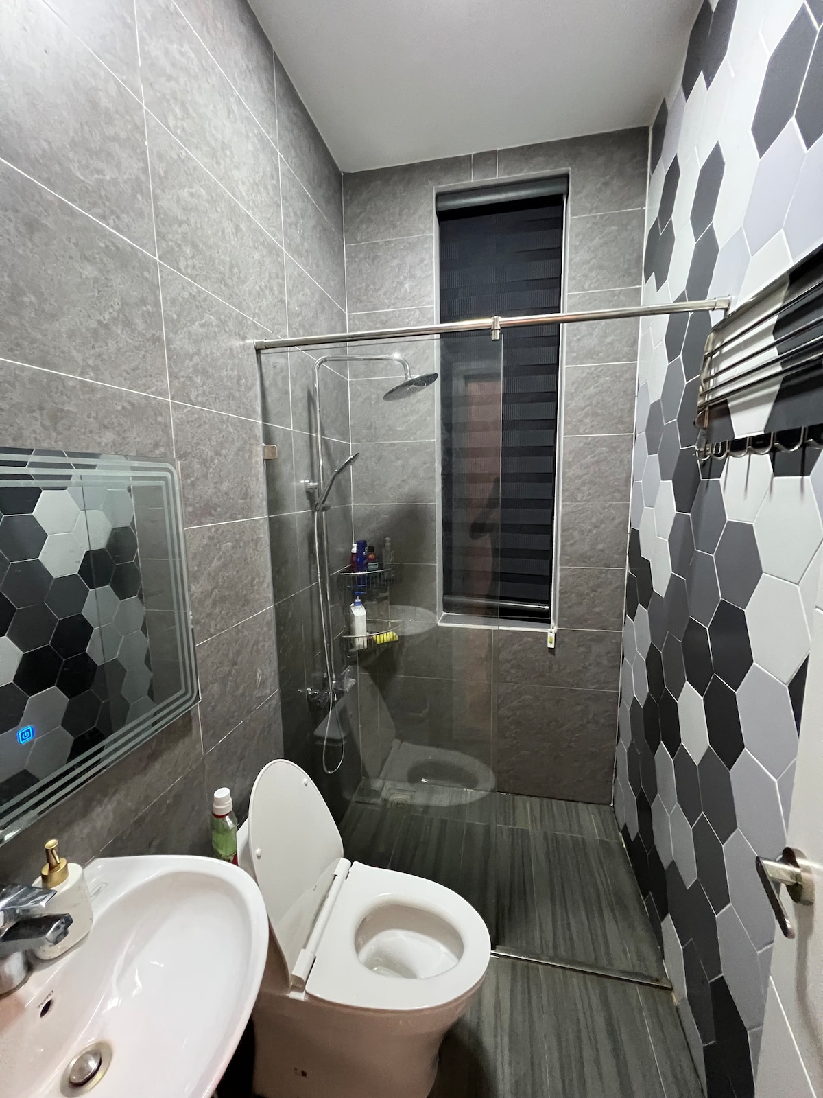 A modern bathroom is presented, featuring sleek gray tile walls and a glass shower enclosure. A white vanity with a round mirror is positioned next to a compact toilet. Dark blinds cover a window, allowing for privacy while natural light is filtered.
