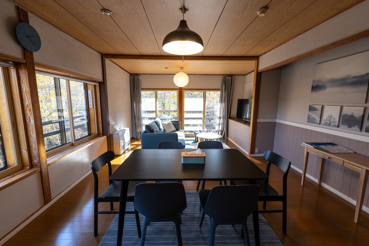 A contemporary dining area features a dark wooden table surrounded by six black chairs. Large windows allow natural light to fill the space, with a cozy seating area visible in the background. Soft furnishings and modern decor enhance the functional layout.