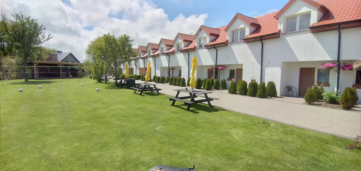 A row of white buildings with red-roofed gables is positioned along a neatly maintained lawn. Picnic tables accompany bright yellow umbrellas, offering shaded seating. A pathway leads toward the structures, with decorative trees and planters enhancing the landscaped environment.