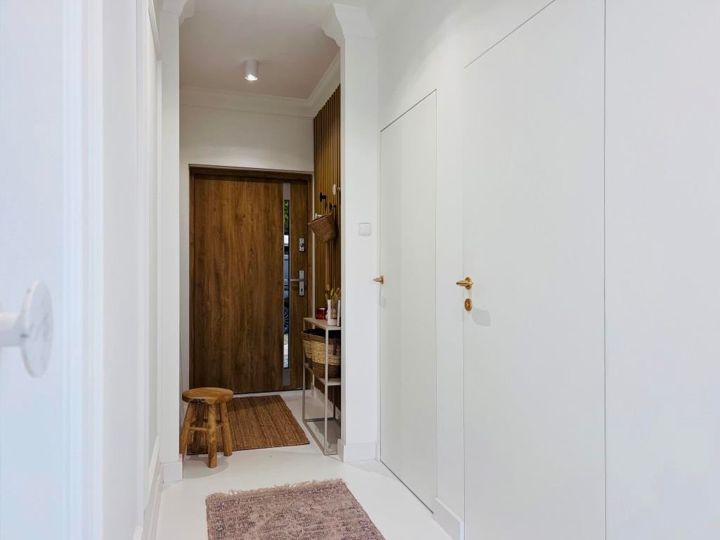 A bright and minimalist hallway is visible, featuring a wooden front door and neutral walls. A small stool is positioned against one wall, while a woven basket and a decorative shelf add functionality. Soft lighting creates an inviting entrance.