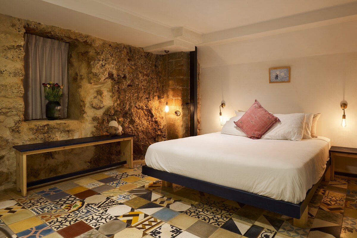 A cozy sleeping area features a spacious bed with a decorative pillow. Natural stone walls provide a rustic backdrop, while pendant lights illuminate the space. A patterned tiled floor adds color, and a bench is placed against a side wall near a window with a sheer curtain.