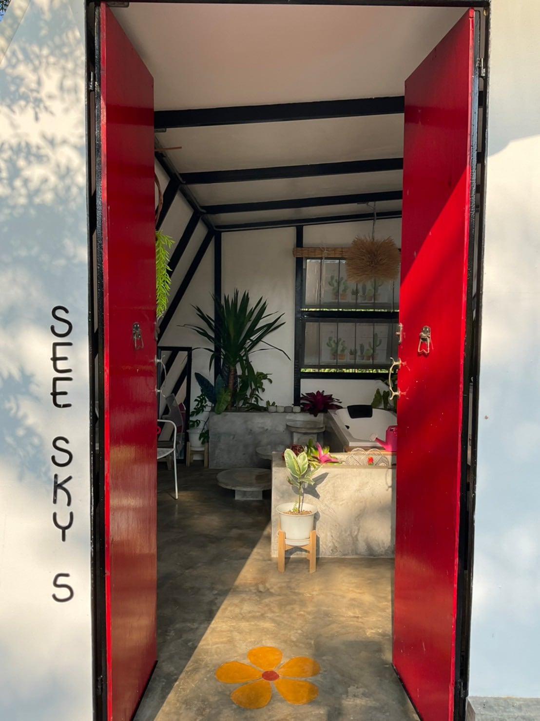 Bright red double doors open into a welcoming entrance, leading to a cozy seating area illuminated by natural light. Green plants and modern decor create an inviting space, with a patterned floor featuring a floral design adding a touch of charm.