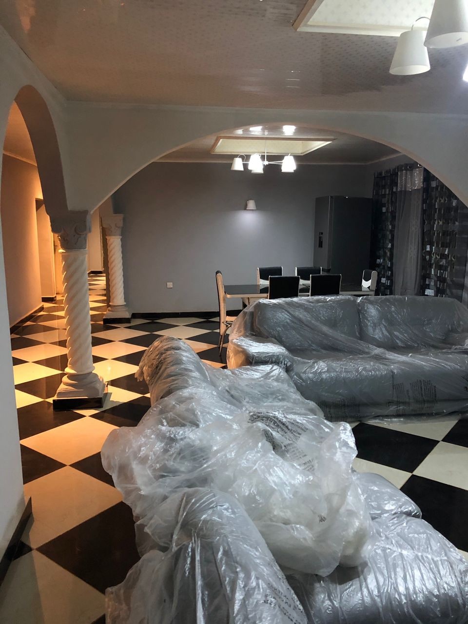 A spacious living area features a striking black and white tiled floor. Wrapped furniture is arranged to create an inviting atmosphere. A dining area with a table and chairs is visible in the background, complemented by modern lighting above.