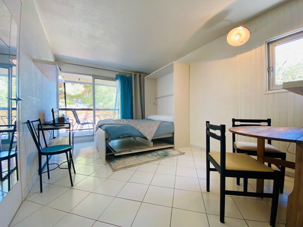 A studio space is featured, showcasing a comfortable bed with a light blanket. A small dining area is visible with a wooden table and four chairs. Natural light enters through a window, and a balcony can be seen in the background, offering a relaxing view.