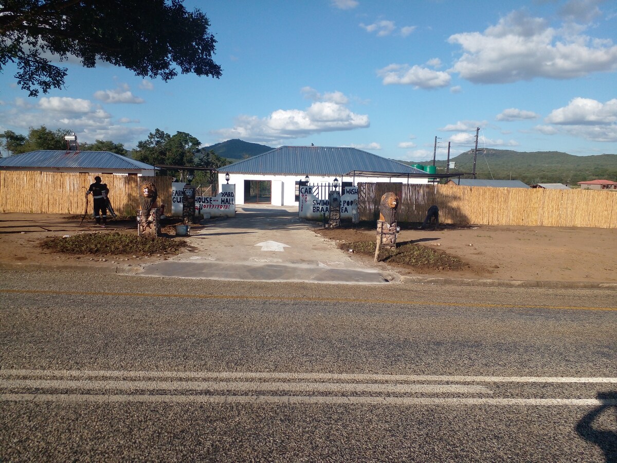 A welcoming entrance to a property is framed by wooden fencing, with two carved figures marking the pathway. A simple building with a metal roof can be seen in the background, surrounded by a scenic landscape featuring mountains and blue skies.