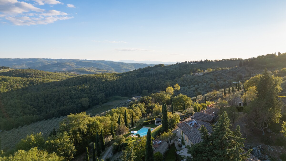An expansive view of rolling green hills stretches towards the horizon, accented by fields of olive trees and cypress trees. In the foreground, a serene swimming pool is visible, surrounded by landscaped gardens that reflect the tranquil beauty of the Chianti countryside.