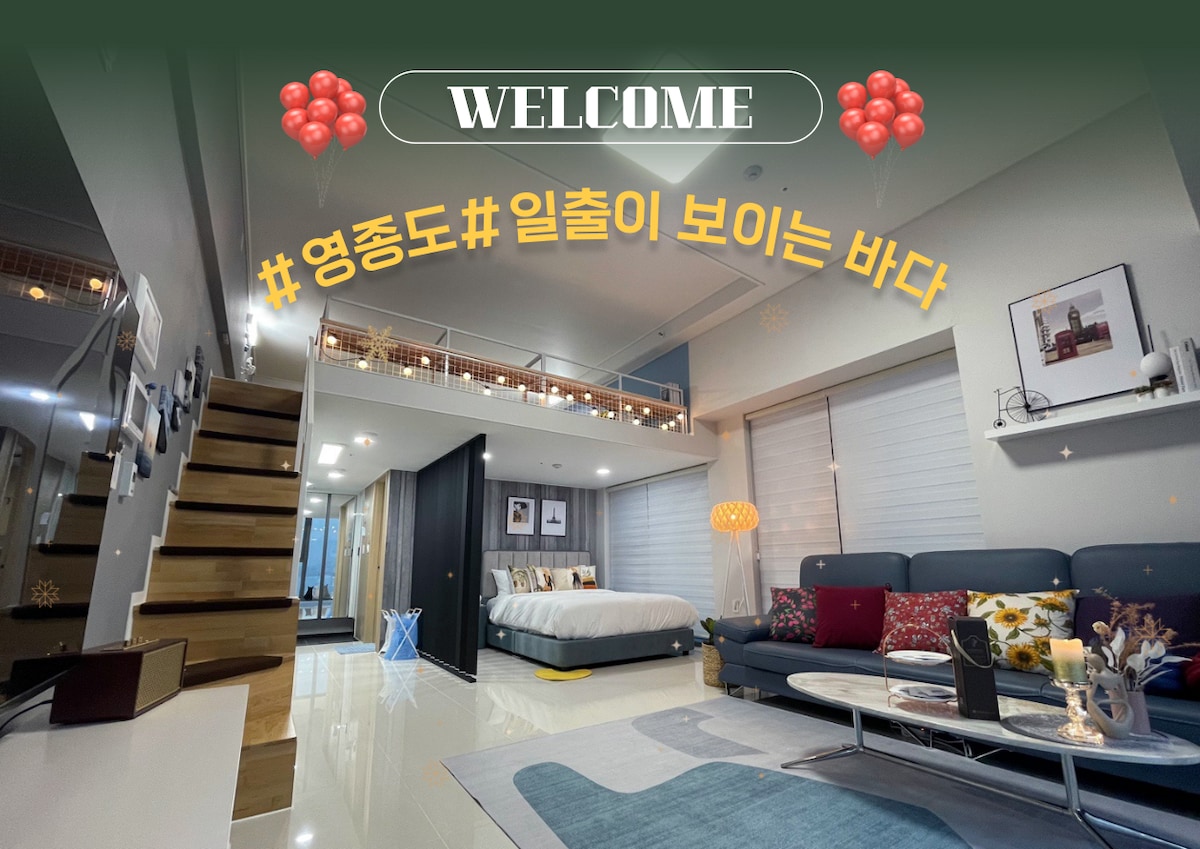 A spacious suite showcases a bright living area with large windows allowing views of the ocean. A comfortable sofa is placed near a coffee table, and a staircase leads to an upper loft. Light-colored walls create an airy atmosphere.