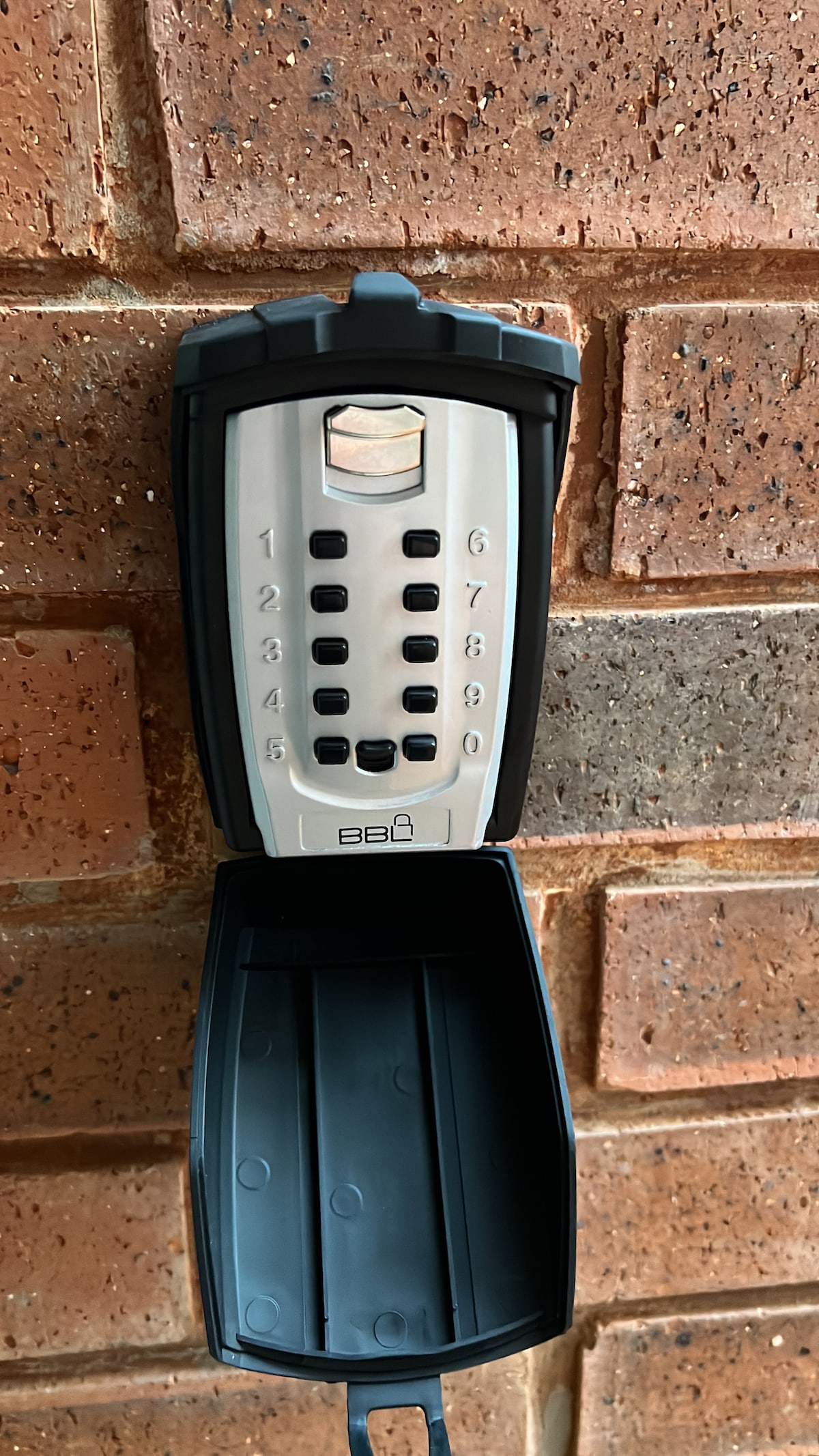A secure combination lockbox is mounted on a brick wall. The box features a numeric keypad with raised buttons, and the lid is open, revealing a compartment for storing a key or access card. The surrounding brick shows a warm, earthy tone.