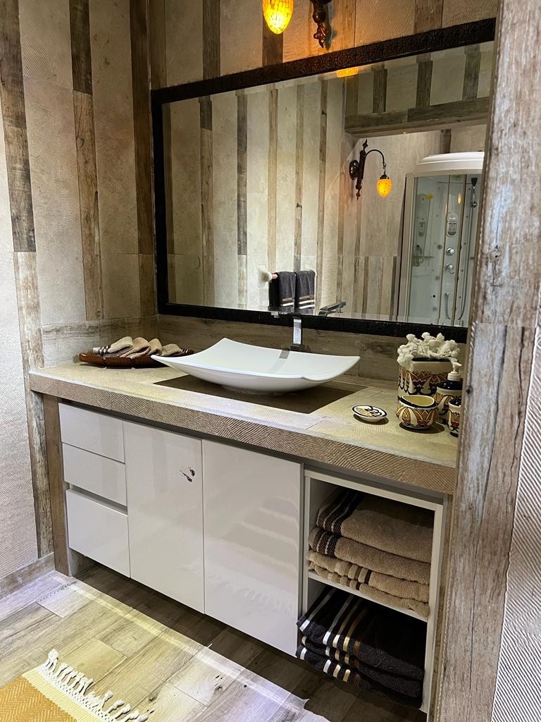 The bathroom features a modern sink atop a stone countertop, complemented by a large mirror framed in rustic wood. Shelves on the side provide neatly arranged towels, while decorative items and toiletries are placed on the countertop, enhancing the organized look.