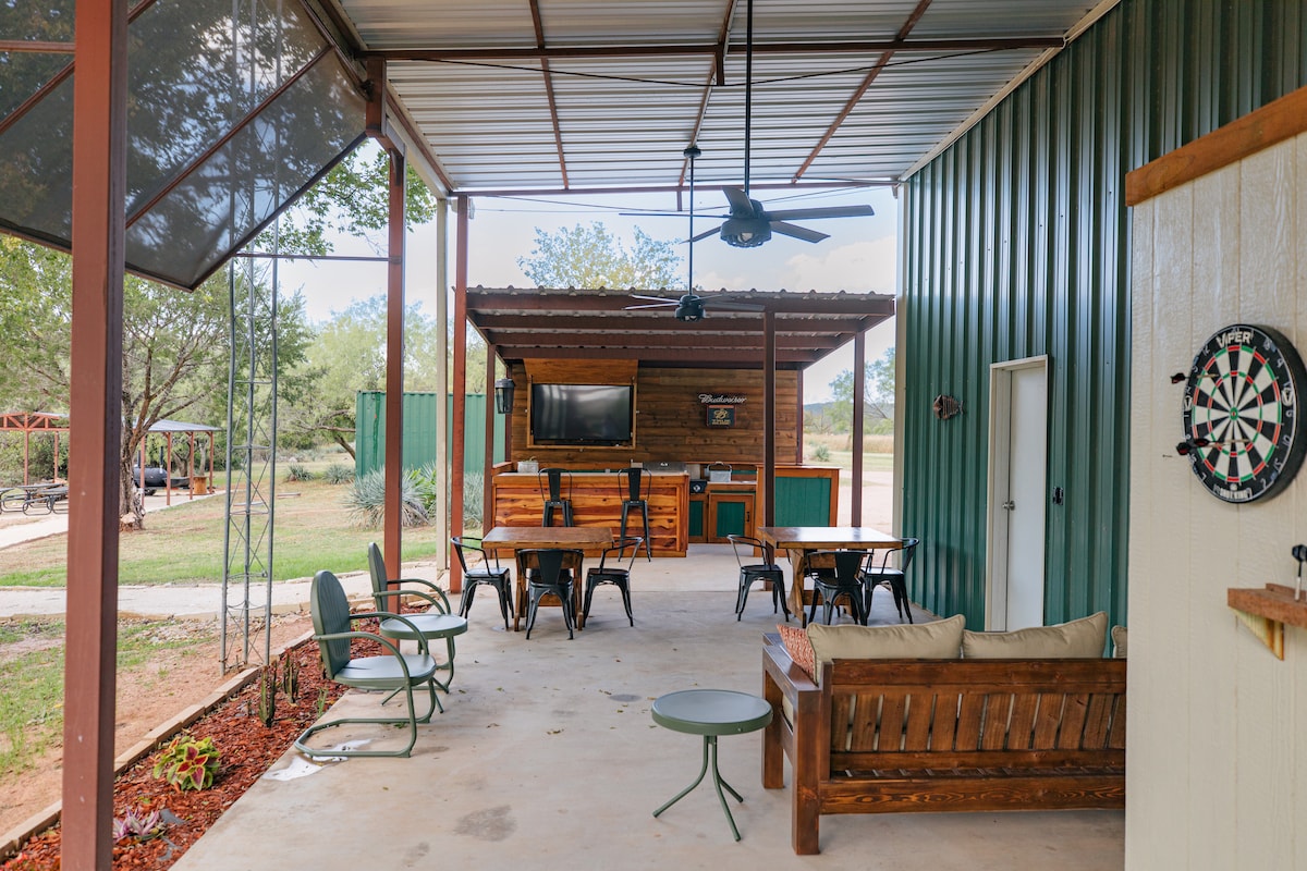 A covered outdoor gathering space features multiple seating options, including a large table and cushioned couches. A dart board is mounted on the wall, while a bar with seating and a television is visible in the background, providing an inviting atmosphere for relaxation and socializing.