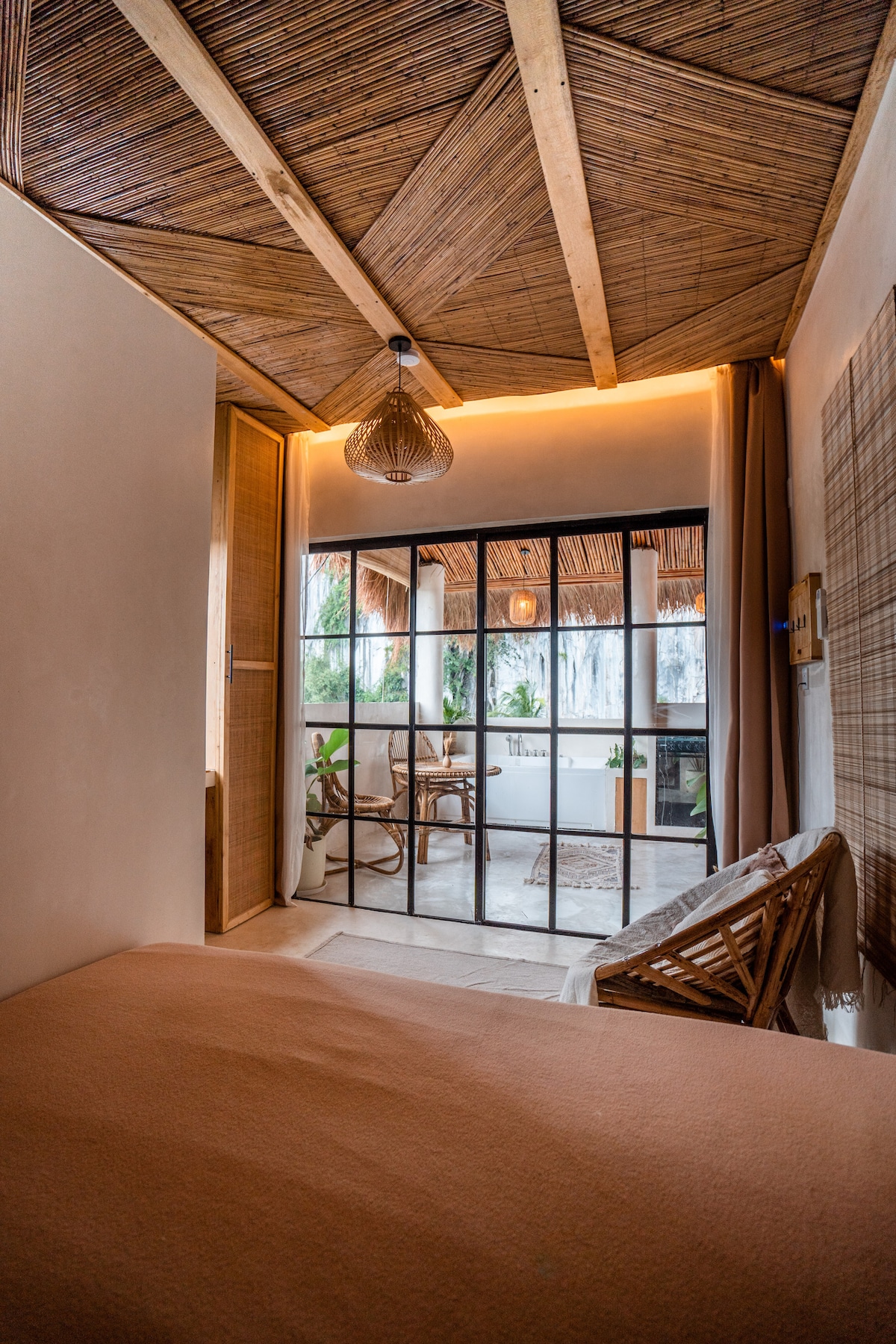 The room features a cozy king-size bed with an earthy-toned duvet, complemented by a rustic ceiling made of natural materials. Glass doors lead to a terrace, allowing natural light to fill the space. Bamboo accents add a touch of warmth to the decor.