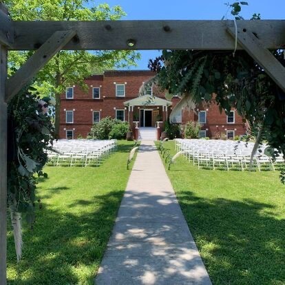 A clear path leads towards a red-brick building, framed by an arch adorned with greenery. Rows of white chairs are neatly arranged on the lawn, inviting guests to gather for an outdoor event. Lush grass surrounds the pathway, creating a serene setting.