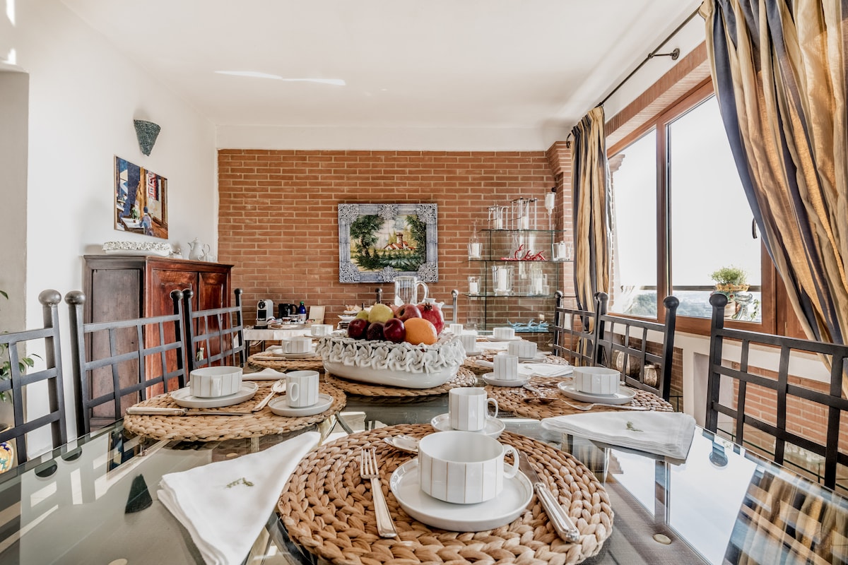 A dining area is presented with a glass table surrounded by six chairs. Each place setting features a white cup and saucer, accompanied by woven placemats. A fruit basket sits centrally, while light filters through the large window, highlighting the warm brick wall and tasteful decor.