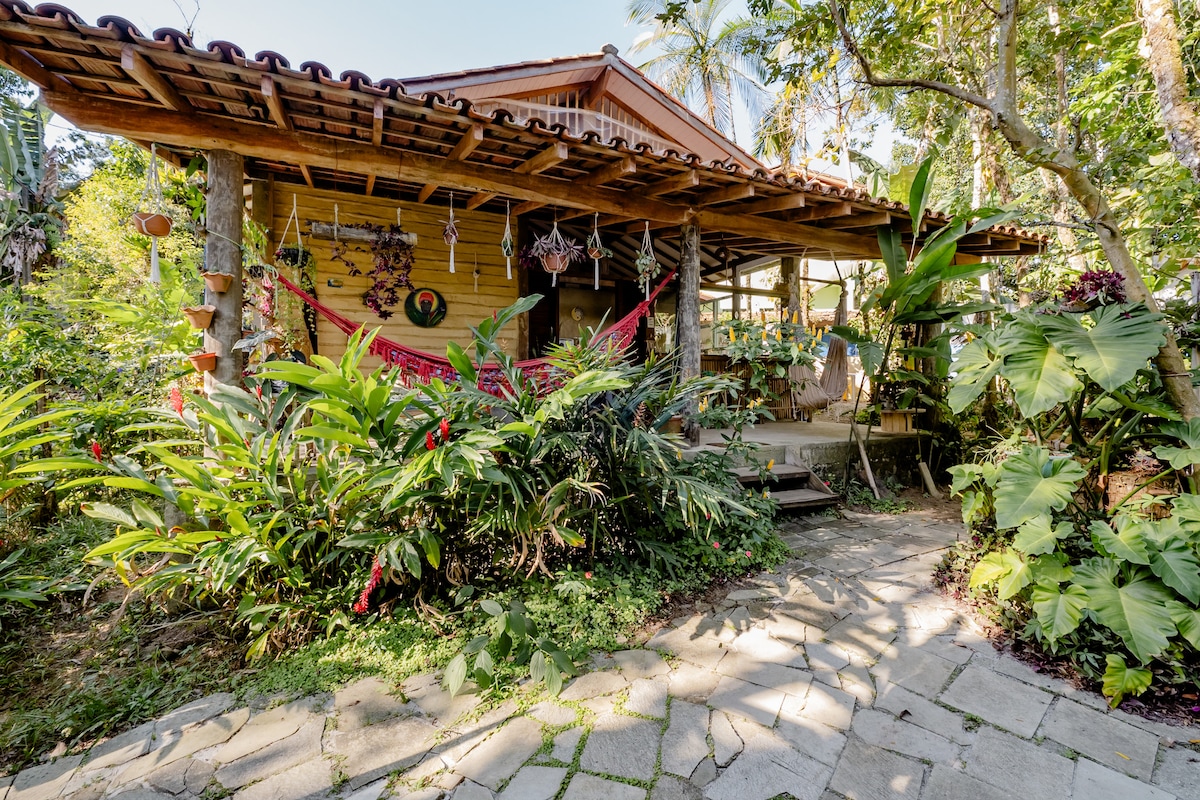 A charming house is surrounded by lush greenery, with tropical plants and flowers framing the entrance. The wooden structure features a covered porch, adorned with colorful hammocks. A stone pathway leads to the front door, inviting guests into the serene environment.