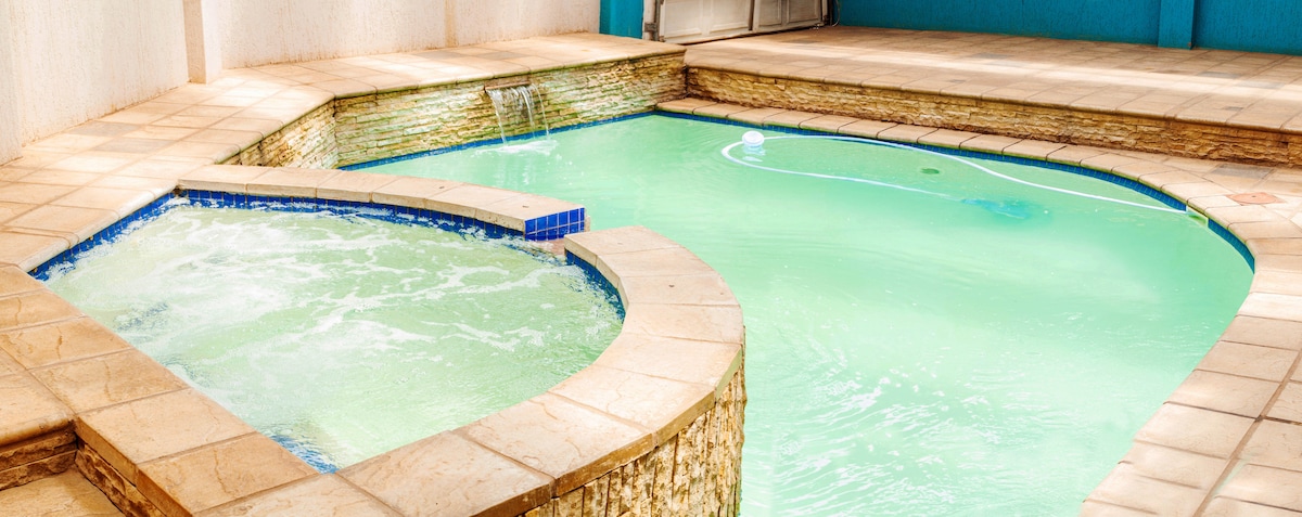 A private outdoor pool area features a gently flowing waterfall and a spacious deck. The pool is designed with a light green water hue, complemented by tiled edges and a jacuzzi section for relaxation. Sunlight casts a warm glow across the serene space.