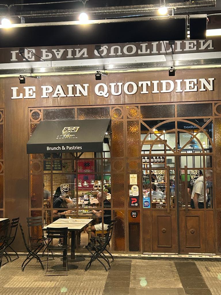 The entrance of Le Pain Quotidien is framed by large glass windows and an inviting wooden facade. A black awning displays the café's name, while outdoor seating is arranged at small tables with metal chairs. Patrons can be seen enjoying their meals inside, illuminated by warm interior lighting.