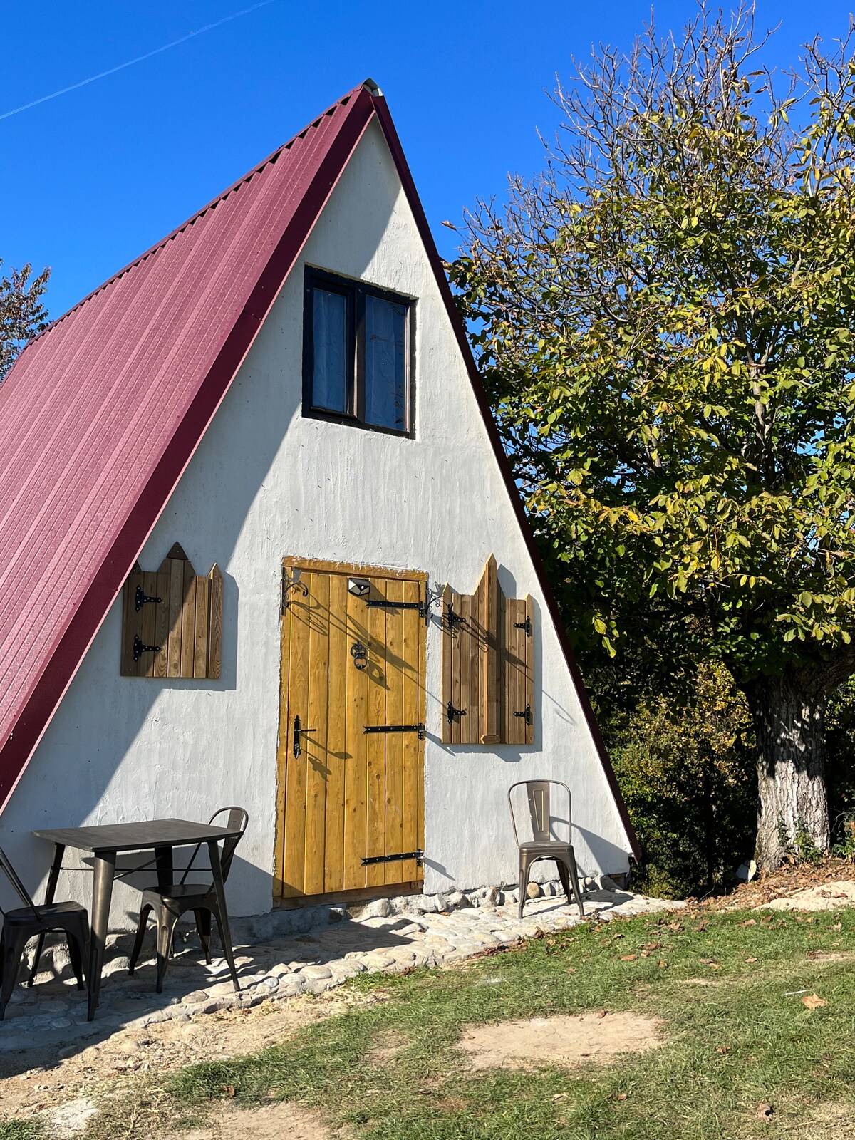 A charming A-frame cabin is set against a clear blue sky, featuring a striking red roof and a textured white exterior. Wooden shutters complement the door and windows, while a small seating area with chairs and a table is situated on the grassy front yard.