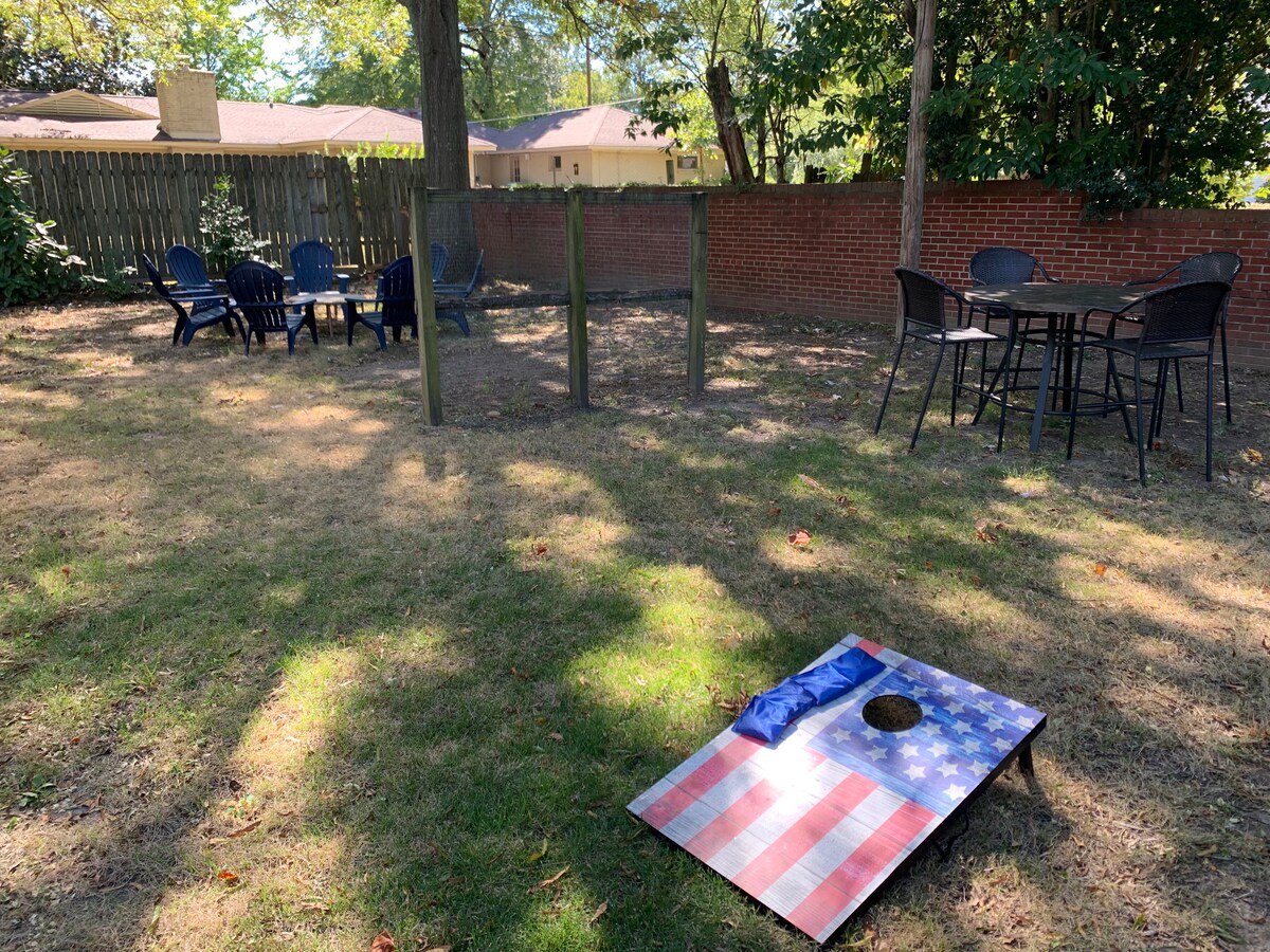 An outdoor space features a grassy area with several seating arrangements, including a set of black chairs around a table. A cornhole board with an American flag design is positioned on the ground, alongside wooden posts that serve as a game target.