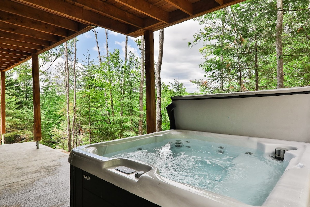 A hot tub is situated on a covered deck, surrounded by a lush forest. Trees are visible in the background, providing a sense of seclusion. The peaceful setting invites relaxation and connects guests with nature.