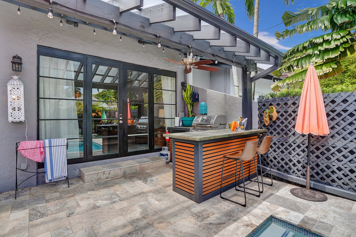 An outdoor kitchen area features a bar with seating and a grilling station. Decorative lighting and lush greenery surround the space, enhancing its inviting atmosphere. A patio umbrella provides shade near the pool, while modern finishes and stone flooring complete the setup for outdoor dining and relaxation.