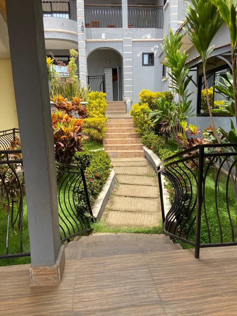 A landscaped pathway leads through vibrant greenery and colorful plants towards the entrance of the lodge. The pathway is flanked by well-maintained gardens, creating a welcoming atmosphere as natural light reflects off the surrounding stone and foliage.