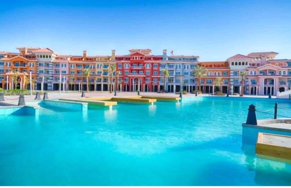 A vibrant resort complex features colorful buildings reflected in a large, clear pool. Palm trees line the edges, and spacious lounging areas are visible, creating a relaxing atmosphere around the water. The sky above is bright and cloudless, enhancing the inviting appearance of the setting.