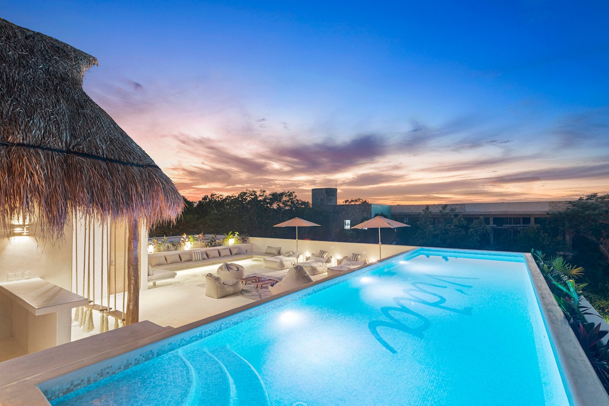 An infinity pool glows in soft blue light, surrounded by lounge areas with large cushions and umbrellas. The sunset casts vibrant hues across the sky, while a thatched-roof structure provides shade nearby. Lush greenery frames the space, creating a peaceful outdoor setting.