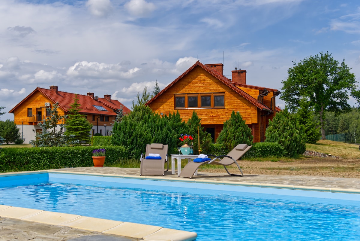 A view of the pool area features a clear blue pool complemented by two lounge chairs positioned nearby. The wooden houses, surrounded by greenery, provide a tranquil backdrop. A small table adorned with a vase of flowers adds a welcoming touch to the outdoor space.