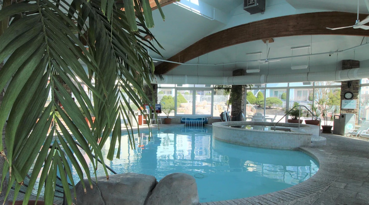 An indoor pool area is revealed, featuring clear water and a spacious layout. Large windows allow natural light to fill the space, while palm-like plants enhance the tropical feel. A hot tub is positioned nearby, creating a relaxing atmosphere for guests.