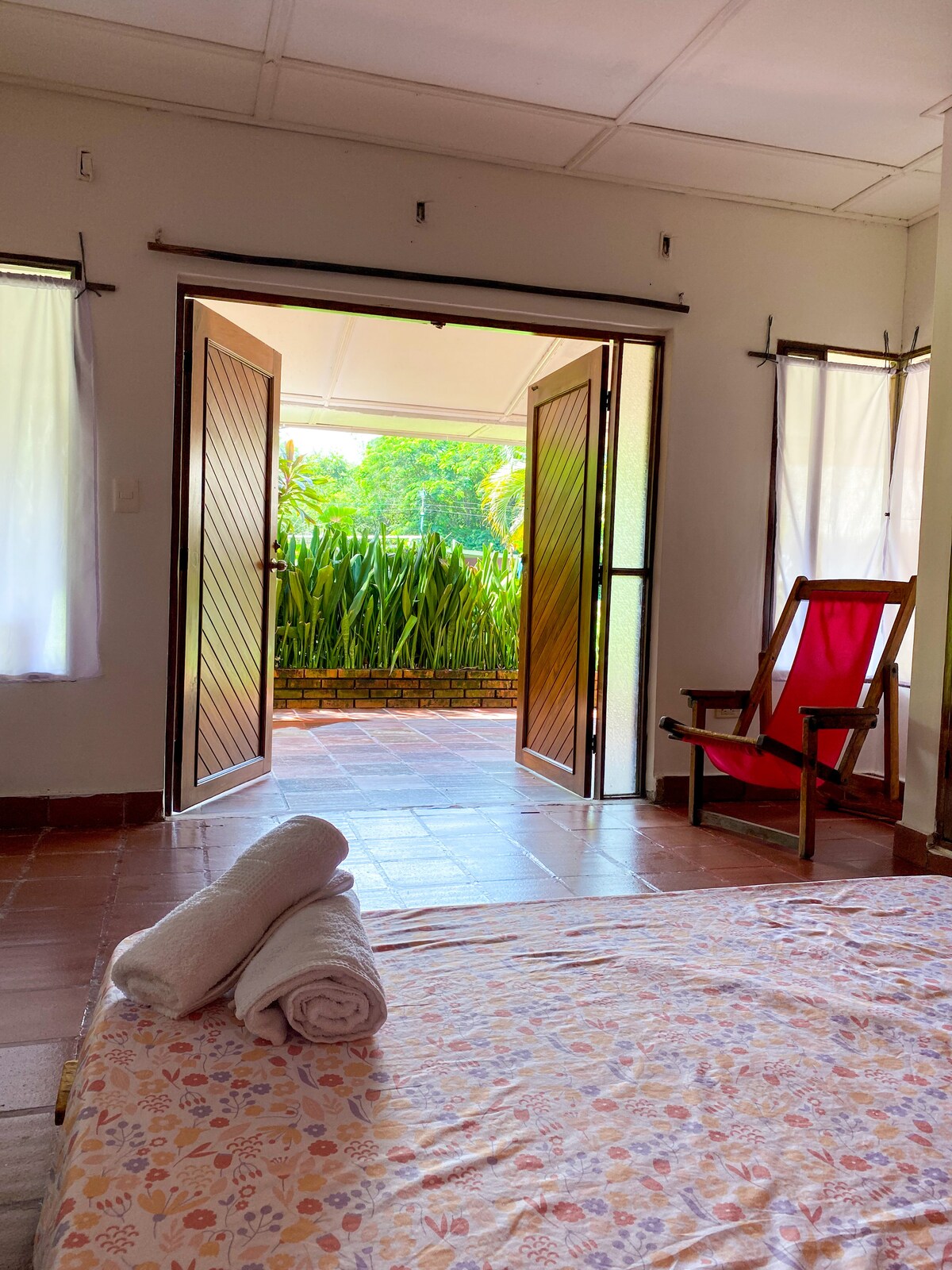Light filters through the open double doors, revealing a view of lush greenery. A spacious bed with neatly arranged towels invites relaxation. A red chair stands nearby, complementing the warm, earthy tones of the room's tiled flooring.